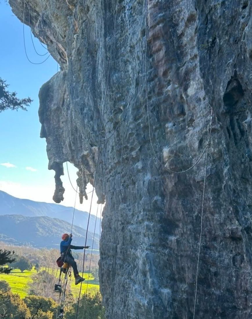 A climber on a rope under an overhanging wall, with green farmland and hills behind