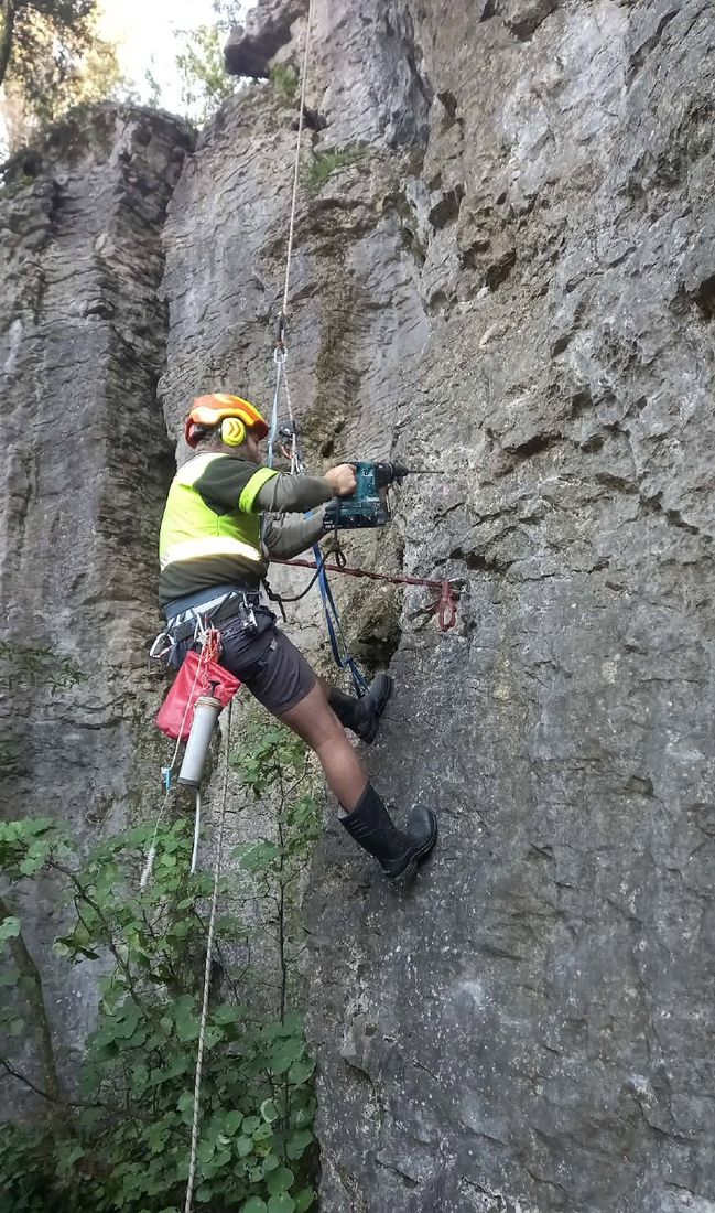 A volunteer in a hi-vis shirt and gumboots drilling on a grey limestone wall